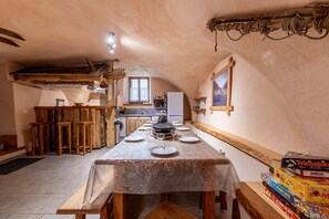 A rustic kitchen with a long dining table set for four, wooden stools, a hanging pot rack, and a window with a view.