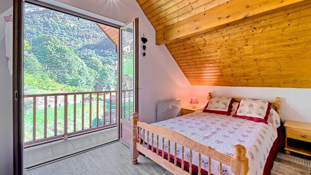 Ein Schlafzimmer mit Holzboden, einem Bett mit gemustertem Überwurf, einem Nachttisch und Blick auf eine grüne Landschaft durch eine Schiebetür.