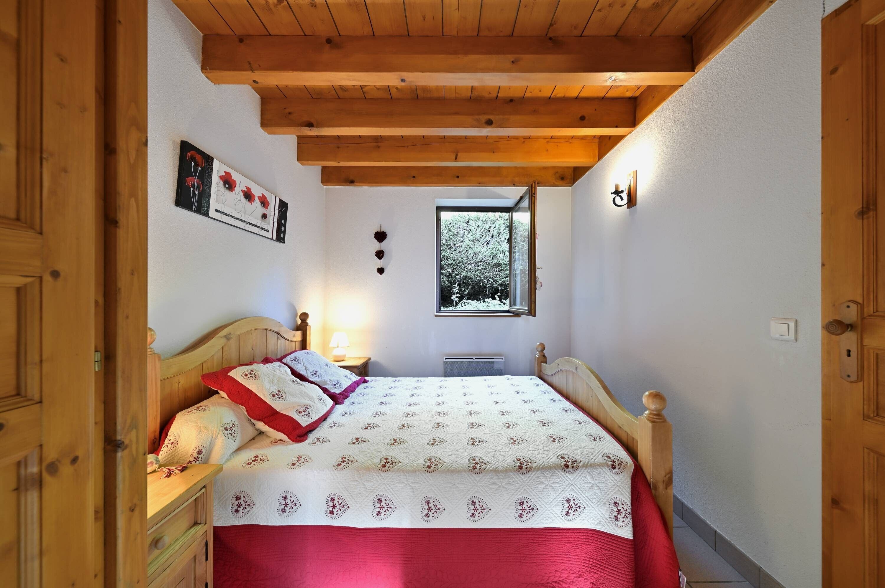A bedroom with a wooden ceiling, a bed with a red and white quilt, a nightstand, a window, and a wall-mounted picture.
