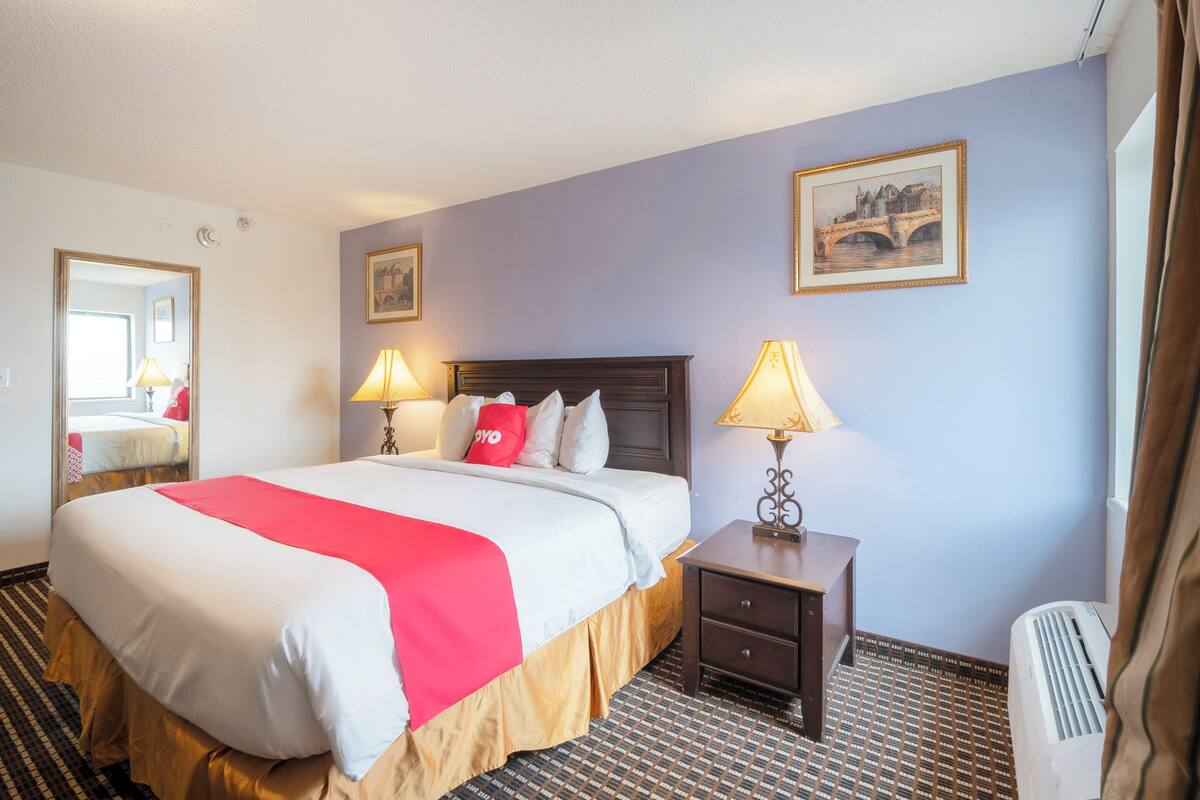 Quarto de hotel com uma cama grande, duas luminárias de cabeceira, mesa de cabeceira, um quadro na parede e janela com cortinas.