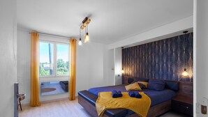 Une chambre moderne avec un grand lit, une fenêtre avec des rideaux, une tête de lit dotée d’un éclairage intégré et un plancher en bois.