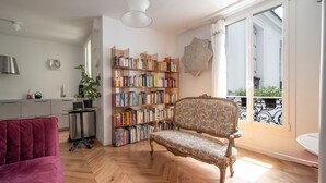 A spacious living area with a herringbone floor, a red sofa, a patterned armchair, a bookshelf, and a balcony with a wrought iron railing.
