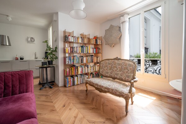 A spacious living area with a herringbone floor, a red sofa, a patterned armchair, a bookshelf, and a balcony with a wrought iron railing. - Appartement Ottomane - Welkeys (Paris)