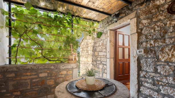 A stone-walled room with a wooden door, a round stone table with a woven placemat, and a potted plant.