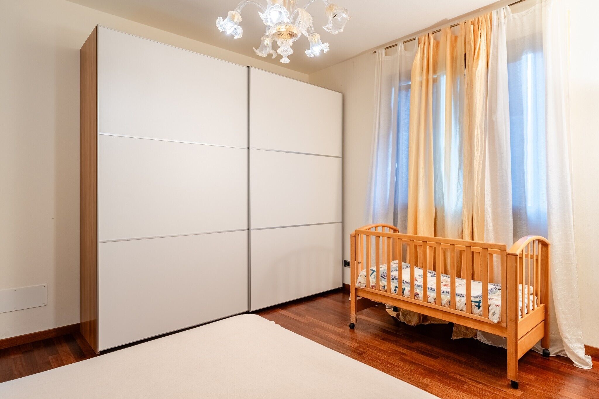 A bedroom with a wooden crib, a large white wardrobe, and a chandelier.