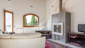 Ein großzügiges Wohnzimmer mit einem weißen Sofa, einem Kamin, einem Fernseher und einem großen Fenster mit Blick auf Bäume.