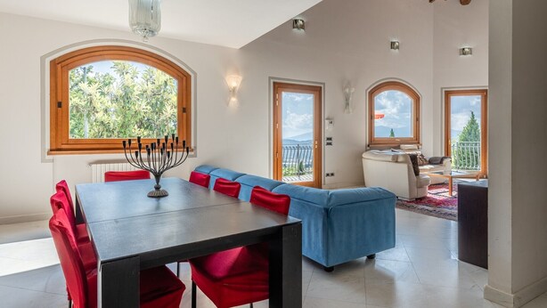 Ein Essbereich mit Tisch, roten Stühlen und einer blauen Couch. Fenster bieten Blick auf Grünflächen.