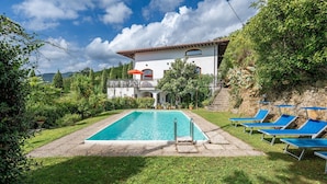 Ein Pool mit Liegestühlen, ein weißes Gebäude mit rotem Dach und eine gebirgige Kulisse.