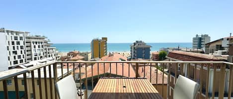 Un balcón con mesa y sillas de madera, con vista a una playa y al skyline de la ciudad.