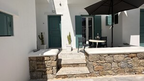 Un bâtiment blanc avec des volets turquoise, un escalier en pierre menant à un patio, et une table avec des chaises placée sous un grand parasol.