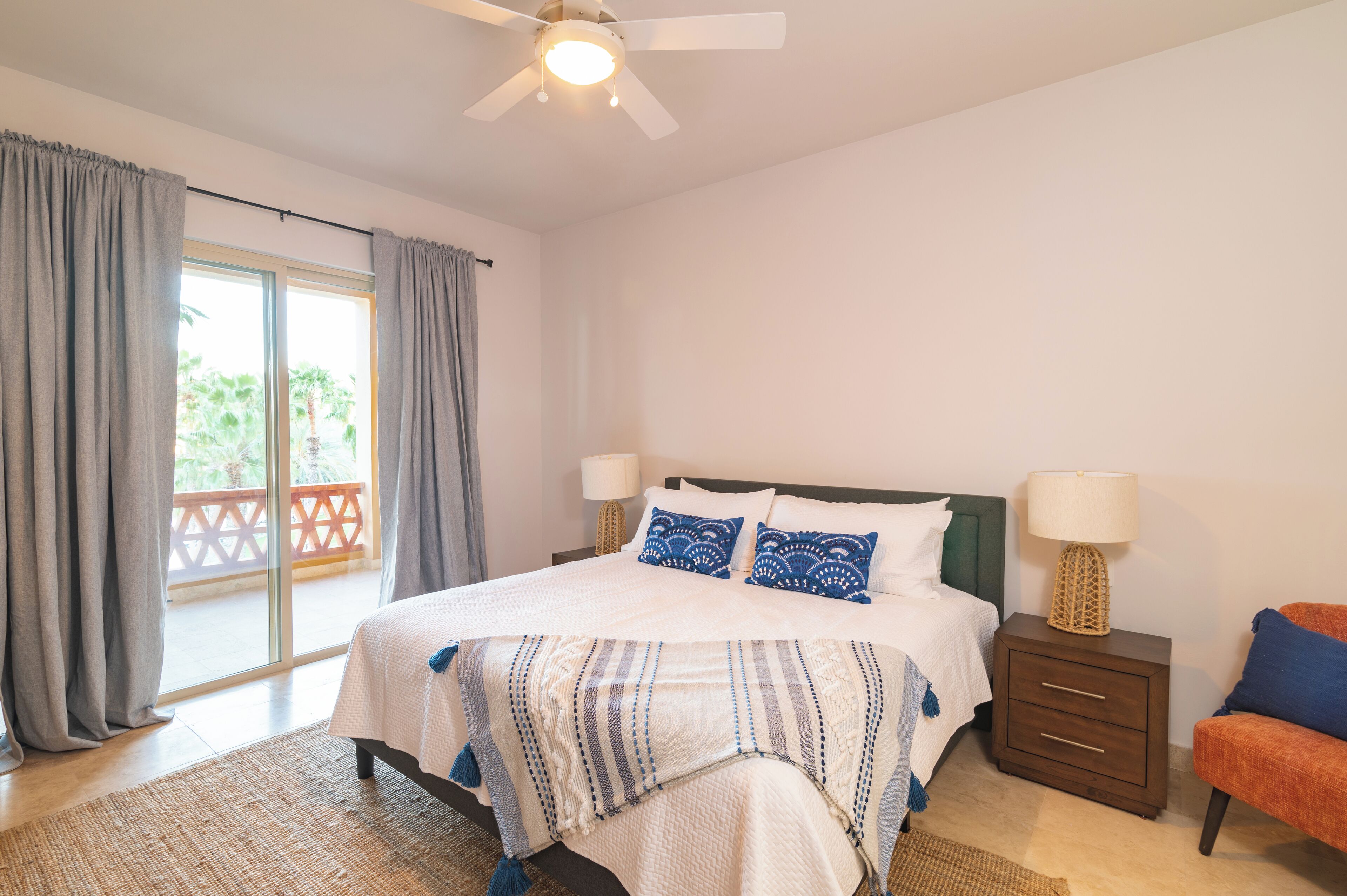 A bedroom with a bed, two bedside tables, a nightstand, a ceiling fan, and a view of a balcony.