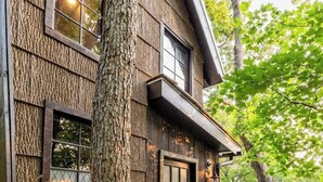 A wooden house with a deck, large windows, and a tree in front.
