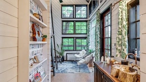 A narrow hallway with a white wall, a wooden floor, a white chair, a wooden cabinet, and a window with a view of greenery.