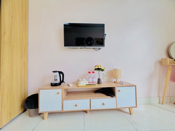 Una habitación con escritorio de madera, televisor montado en la pared, tetera, lámpara y una mesita con botellas y un jarrón de flores.