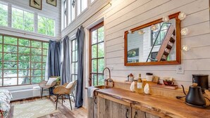 Una cocina con encimera de madera, un lavabo y un espejo grande. Hay ventanas con cortinas, una silla y un radiador.