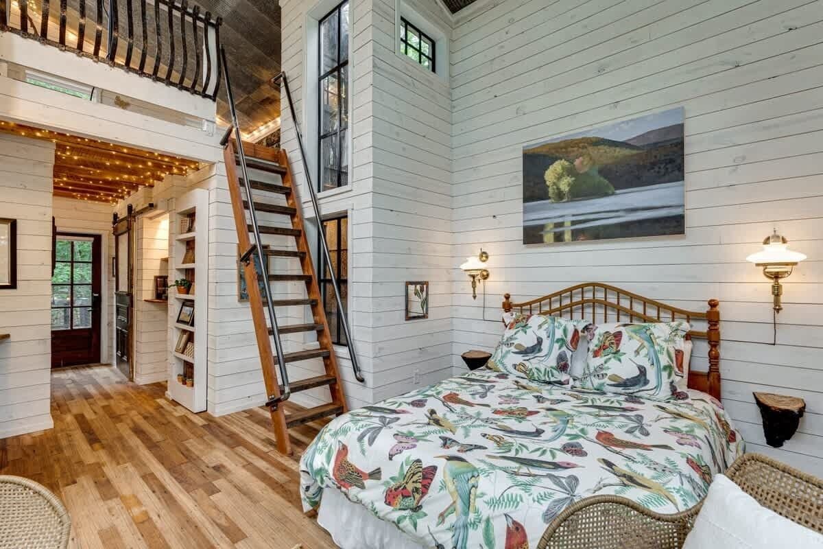 A bedroom with a bed, a ladder, a wooden staircase, a painting, and wall sconces.