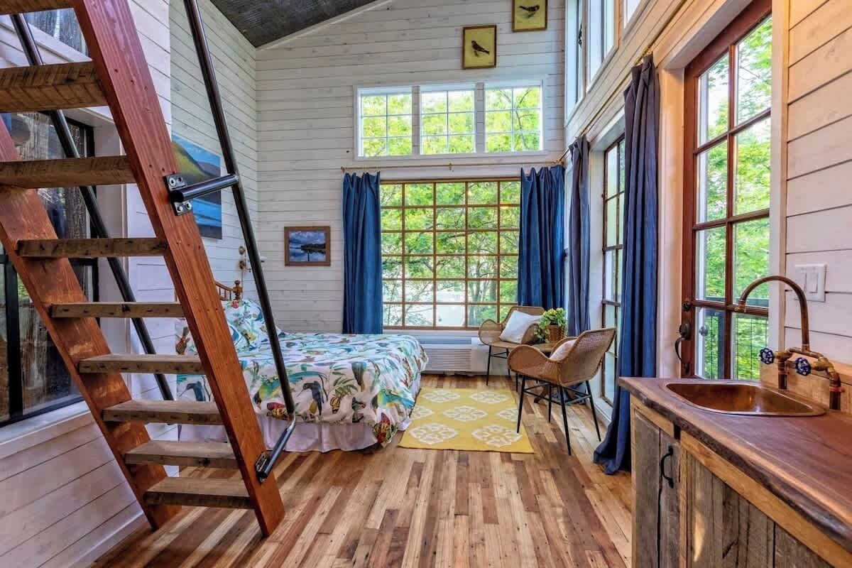 Une pièce avec une échelle en bois, un lit recouvert d’une courtepointe aux couleurs vives, une fenêtre avec des rideaux bleus, un bureau en bois et un lavabo avec un robinet en cuivre.