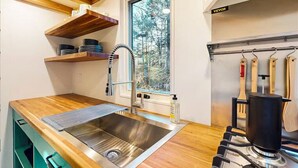 Une cuisine dotée d’un évier en acier inoxydable, de plans de travail en bois, d’une fenêtre donnant sur les arbres et d’une étagère suspendue pour ranger les ustensiles.