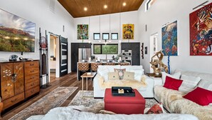 A spacious living area with a large sofa, a red ottoman, and a dining table. There is a kitchen area with modern appliances and a dining table. The room has a high ceiling with wooden beams and artwork on the walls. - Beautiful Views Of Cathedral Rock, Hot Tub, Fire Pit, Wildlife Sightings, And Stargazing (Sedona)