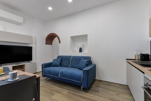 Un espacio habitable moderno con un televisor de pantalla plana, un sofá azul, una zona de cocina y suelo de madera.