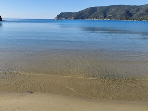 A beach with clear shallow waters, a sandy shore, and a distant mountain range.