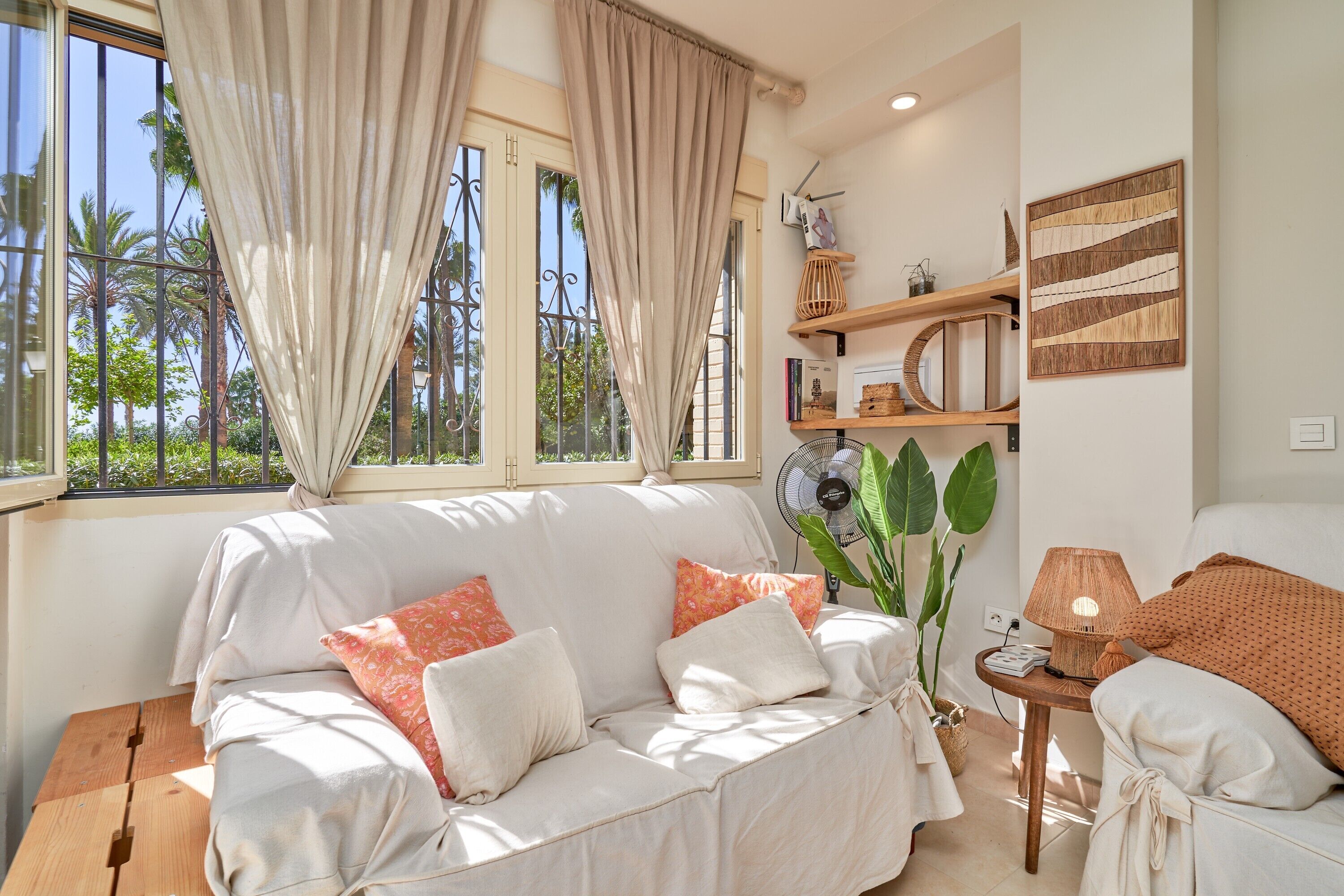 A room with a sofa, a lamp, a fan, and a window with curtains.