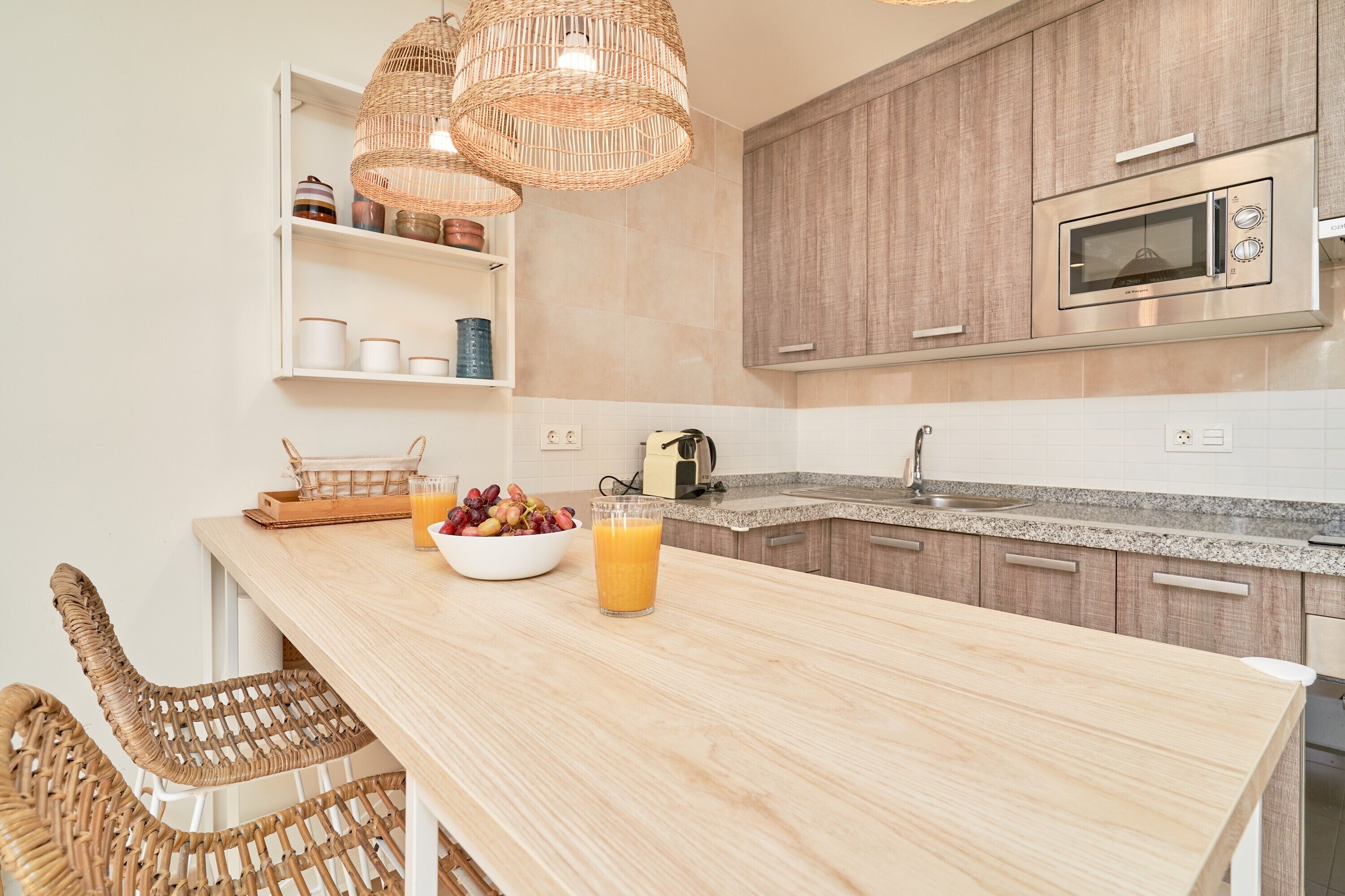 A kitchen with a wooden island, wicker chairs, a fruit bowl, a glass of juice, and a microwave.