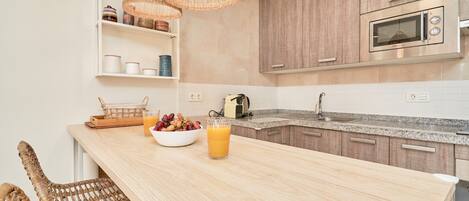 A kitchen with a wooden island, wicker chairs, a fruit bowl, a glass of juice, and a microwave.