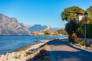 A coastal road with a stone wall on one side and a view of a town with a tower and mountains in the background. - Lake Front Villa Schlosser (Malcesine)