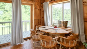 A wooden cabin interior with a round table and four chairs, large windows with curtains, and a view of greenery outside. - The Rustic Retreat: Elegant cabin with WiFi & AC in an enchanting location! (Pearl City)