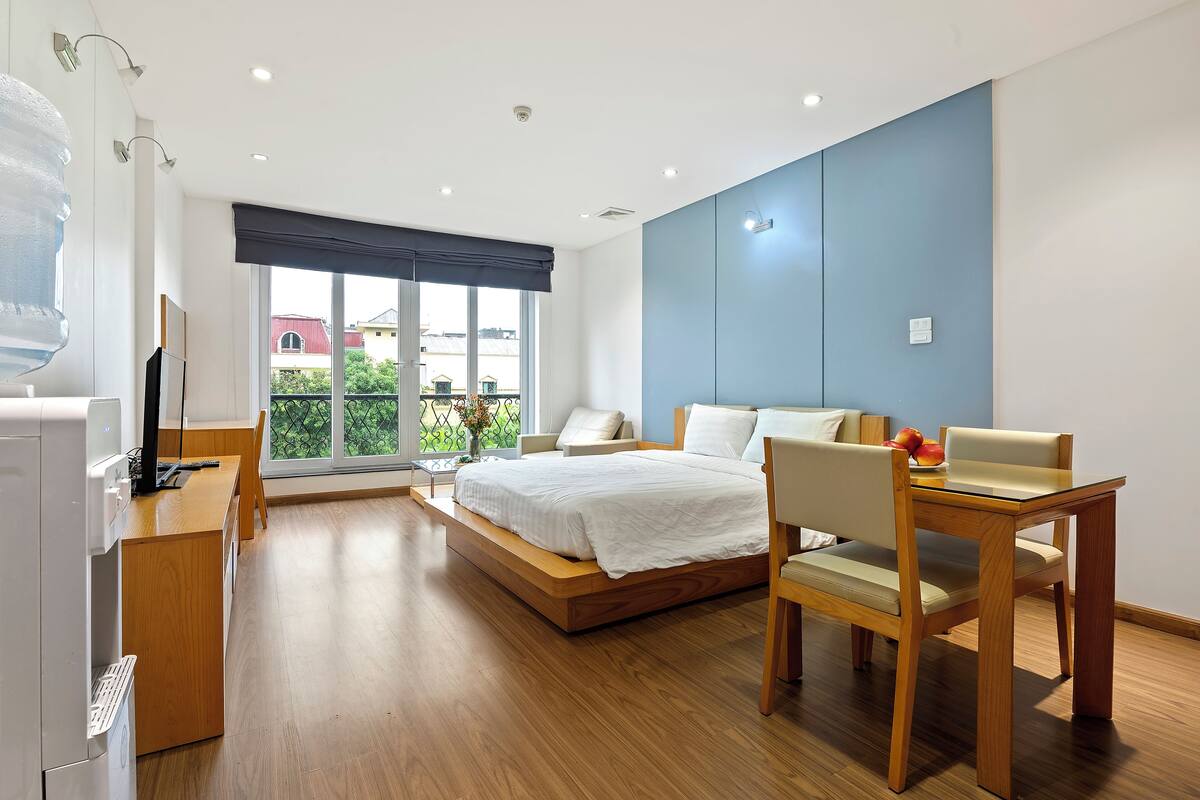 Una habitación de hotel moderna con cama, escritorio, silla y un amplio ventanal con vistas a zonas verdes y edificios.