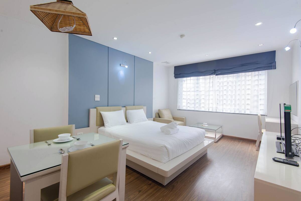 Una habitación de hotel moderna con una cama, un escritorio con silla, una mesita pequeña y un ventanal con persianas.