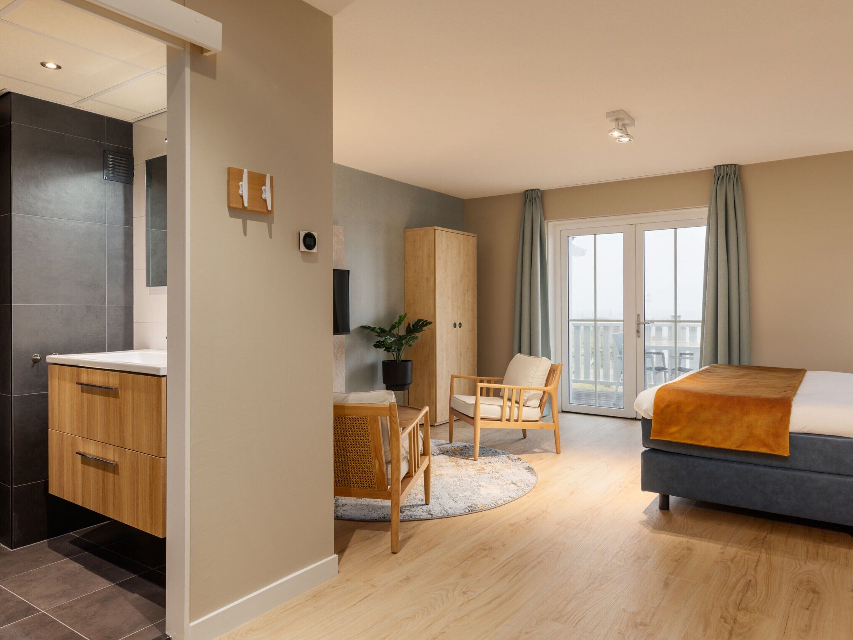 Une chambre d’hôtel moderne comprenant un lit, une armoire en bois, un coin salon avec une chaise et une petite table, ainsi qu’une grande fenêtre avec des rideaux.