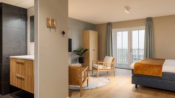 Une chambre d’hôtel moderne comprenant un lit, une armoire en bois, un coin salon avec une chaise et une petite table, ainsi qu’une grande fenêtre avec des rideaux.