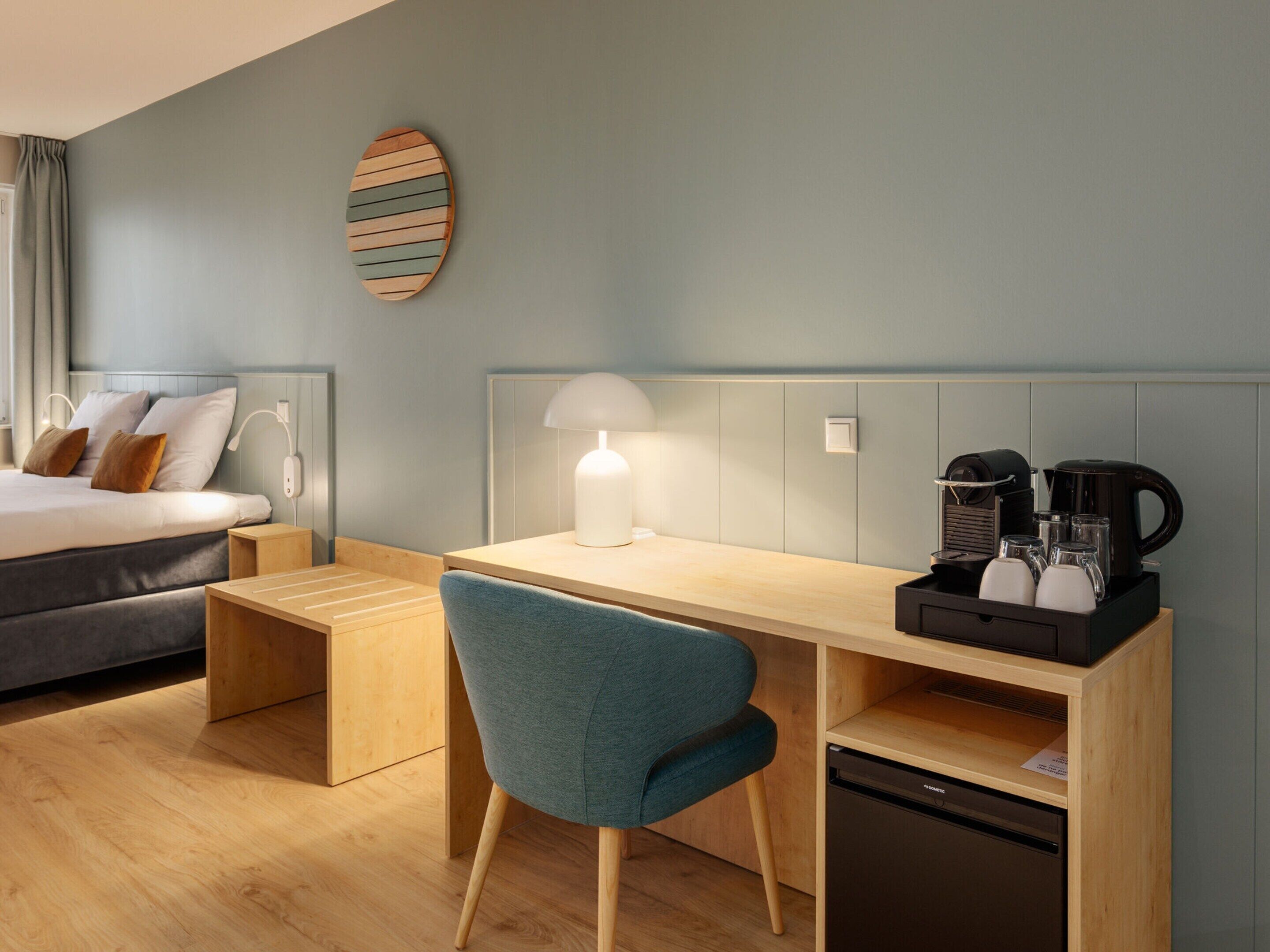 A modern hotel room with a bed, a desk with a lamp, a coffee maker, and a dishwasher.