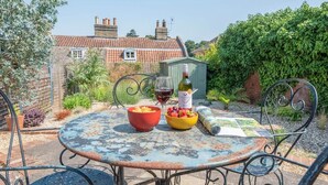 A round metal table with a rustic design, surrounded by metal chairs. A bottle of wine and bowls of fruit are placed on the table.