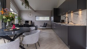 A modern kitchen with dark cabinetry, a marble countertop, and a built-in stove. There is a dining area with a round table and grey chairs. - Eco Villa Near Forest in Netherlands (Oirschot)
