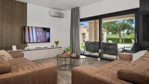 Een moderne woonkamer met een flatscreen televisie, twee bruine banken, een kleine ronde tafel en een glazen schuifdeur die leidt naar een buitenruimte met een zwembad en tuinmeubilair.