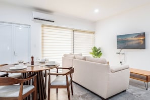 A modern living room with a sofa, dining table, chairs, and a TV mounted on the wall.