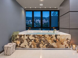 Ein modernes Badezimmer mit einer großen Badewanne, Blick auf eine Stadtkulisse durch große Fenster und einer Marmorarbeitsplatte.