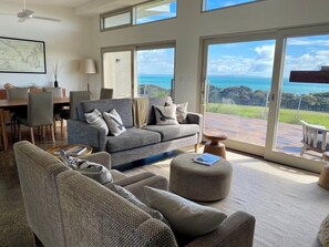 A spacious living room with a grey sofa, a round ottoman, and a dining area with a wooden table and chairs. Large windows offer a view of the ocean and greenery. - Oceanview Eco Villas (Nepean Bay)