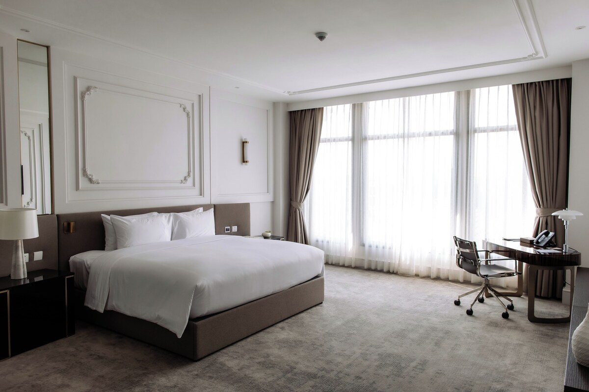Une chambre d’hôtel avec un grand lit, un bureau avec une chaise et une fenêtre avec des rideaux.
