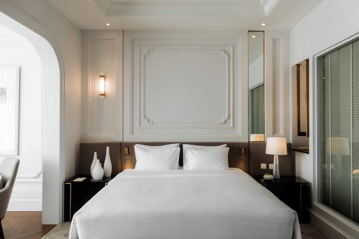 Een hotelkamer met een groot bed, twee nachtkastjes, een stoel en een groot raam met luxaflex.