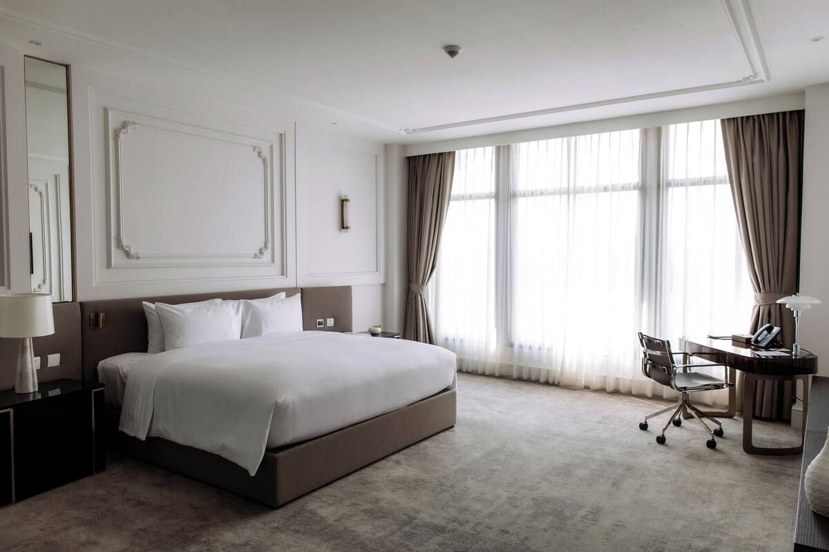Een hotelkamer met een groot bed, een bureau met stoel, een lamp en een raam met gordijnen.
