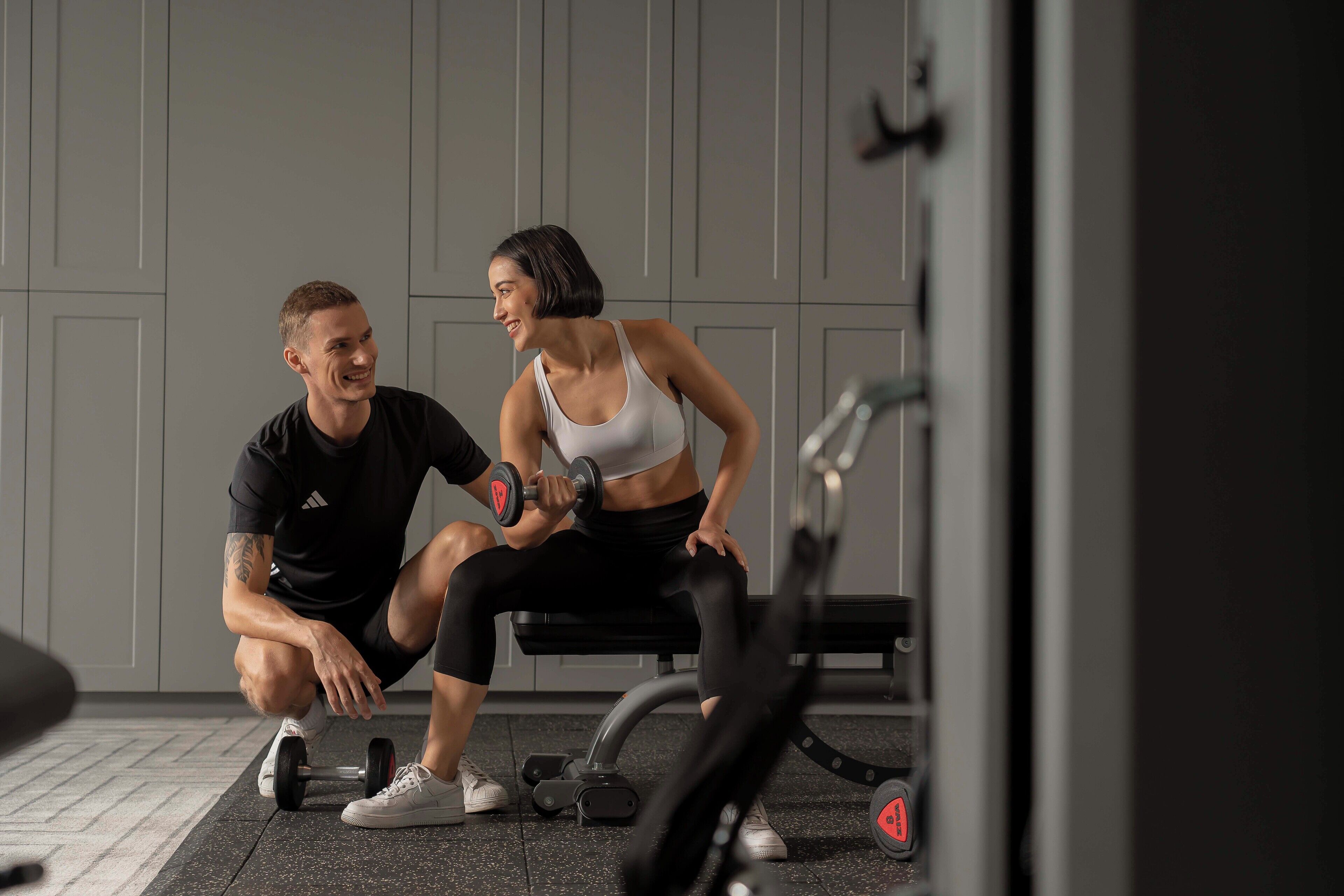 Two people exercising with dumbbells in a gym with grey walls and a bench.