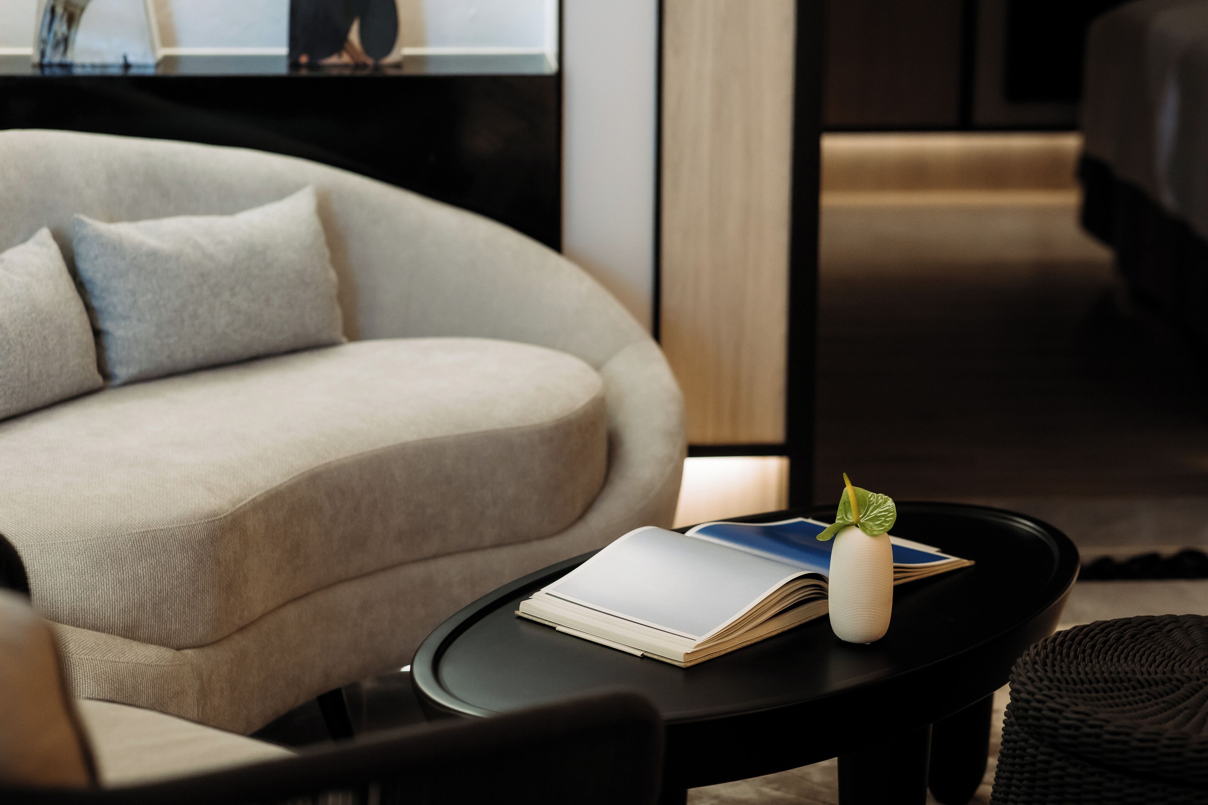 A modern living room with a sofa, a small round table with a book and a decorative vase, and a television in the background.
