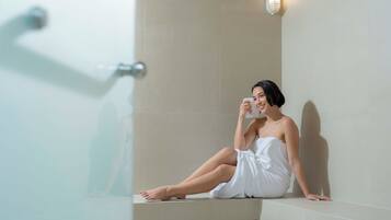 A person sitting in a bathtub with a towel wrapped around their body, a showerhead, and a wall-mounted light fixture.