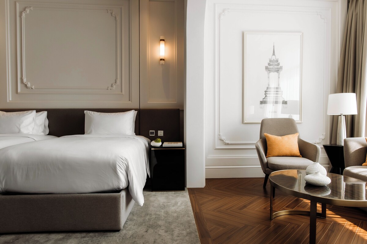 A hotel room with a bed, a chair, a table, and a wall-mounted lamp.