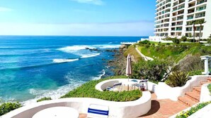 Un balcon avec une table blanche et des chaises bleues, donnant sur une plage où l’on peut voir des vagues et, au loin, un hôtel.
