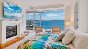 Une chambre avec un lit, une télévision, une cheminée, un balcon avec des chaises et une vue sur la mer.